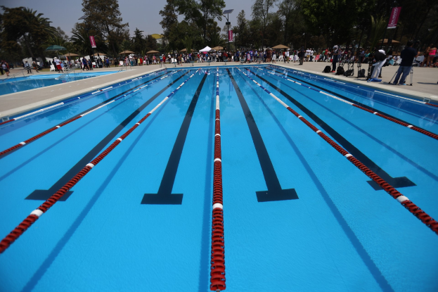 Gro├čer Schwimmbad mit Bahnentrennungen, umgeben von Menschen mit Taschen und Gegenst├Ąnden, mit Zelten, Transparenten, Masten, einem Schild und einem bew├Âlktem Himmel im Hintergrund.