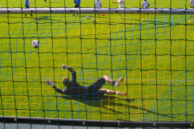 Ein Fussballer wehrt einen Ball vor einem Tor ab, mit einem Netz im Vordergrund und einigen Menschen im Hintergrund, während mehrere Bälle auf dem Boden verstreut sind.