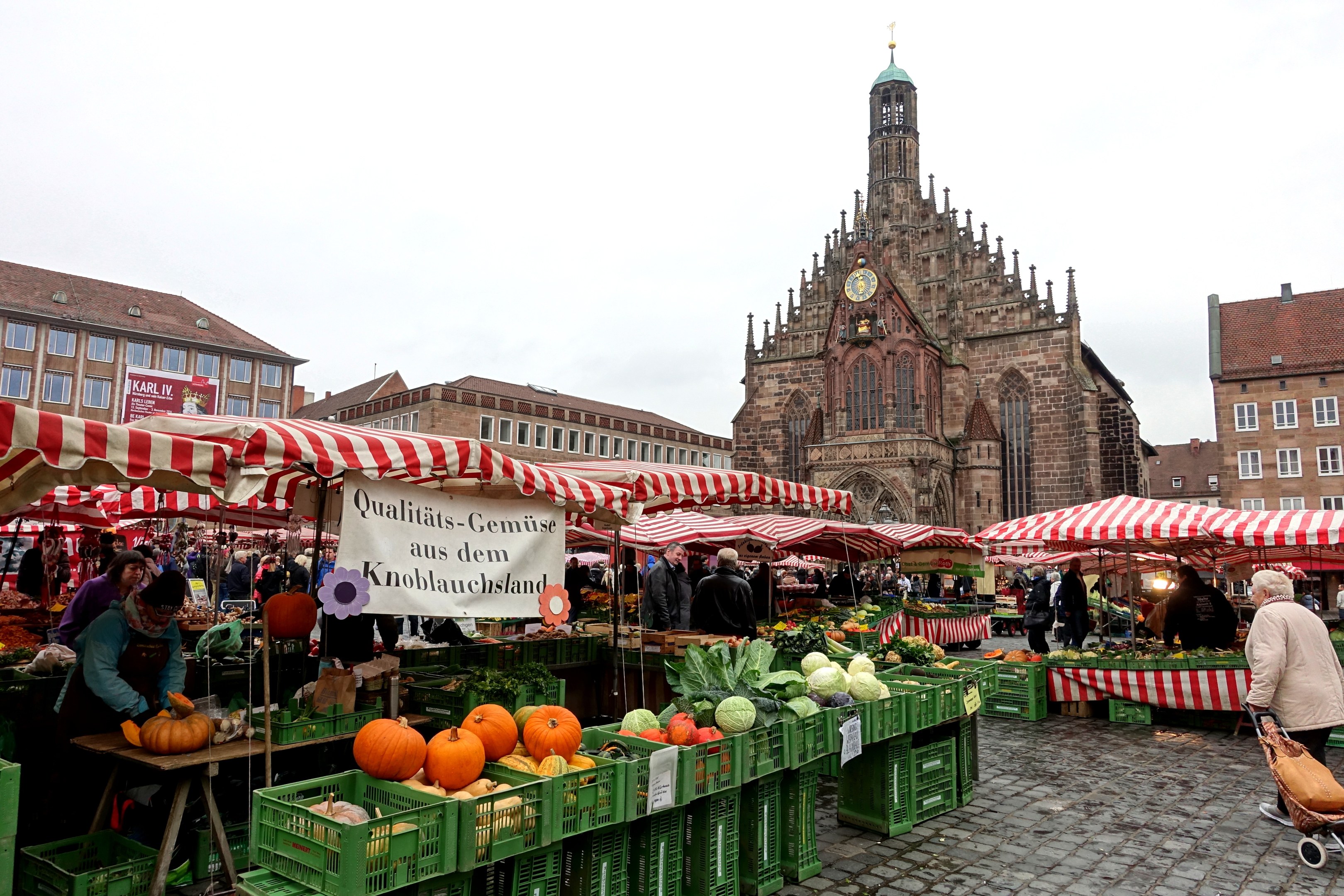 Ein belebter Markt in Nürnberg, Deutschland, mit farbenfrohen Obst- und Gemüseauslagen, Menschen mit Taschen und aufgestellten Zelten im Hintergrund, mit Gebäuden und einem Uhrenturm.