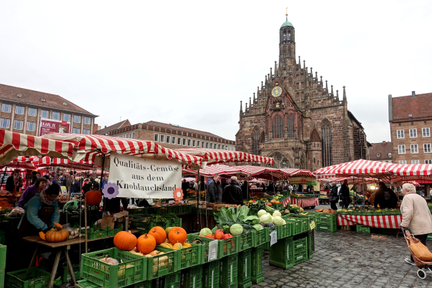 Ein belebter Markt in Nürnberg, Deutschland, mit farbenfrohen Obst- und Gemüseauslagen, Menschen mit Taschen und aufgestellten Zelten im Hintergrund, mit Gebäuden und einem Uhrenturm.