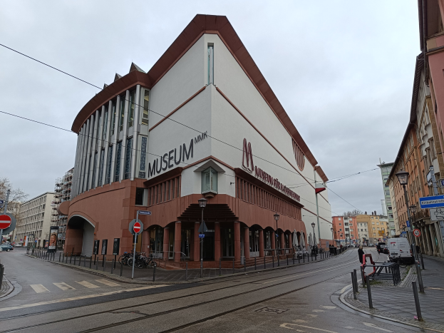 Großes Gebäude mit vielen Fenstern, identifiziert als Museum of Modern Art in München, Deutschland, umgeben von Straßeninfrastruktur und Passanten, mit Bäumen und bewölktem Himmel im Hintergrund.