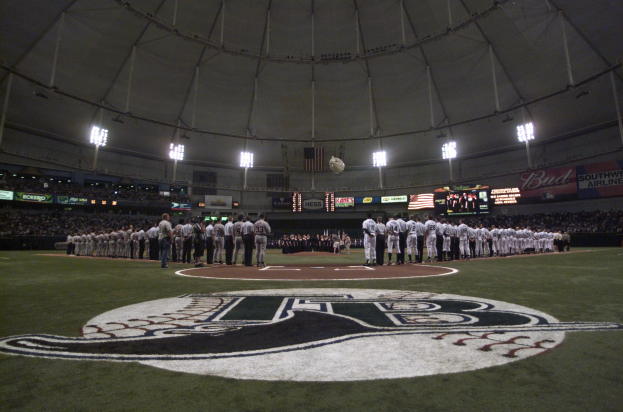 Baseballspieler auf einem Feld mit Fans in den Tribünen, Logo auf dem Boden und Stadionbeschilderung, Bildschirme, Fahnen und eine Kuppelstruktur im Hintergrund.