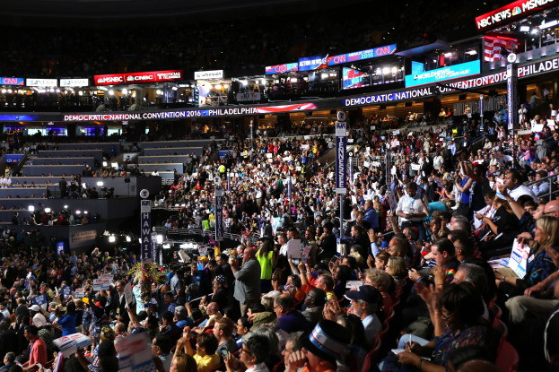 Große Menschenmenge vor einer hell erleuchteten Bühne auf der Democratic National Convention 2016 in Philadelphia, viele halten Schilder, mit Treppen zur Bühne und Texttafeln drumherum.