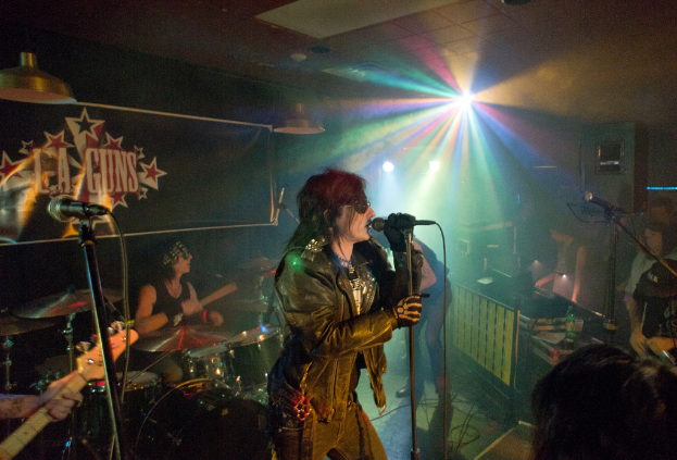 Eine Frau in einer schwarzen Jacke singt in ein Mikrofon auf der Bühne, mit einem Mann in Sonnenbrille und einem Headband, der ein Instrument spielt, und einem Plakat mit 'LA Guns' im Hintergrund.