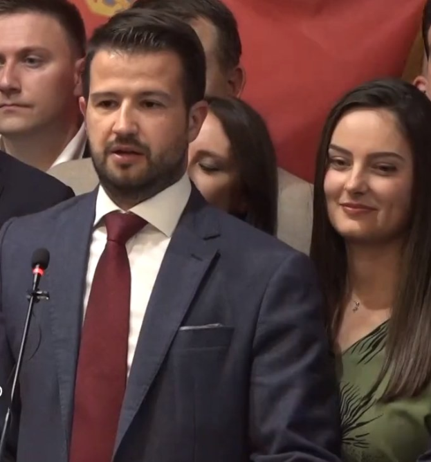 Ein Mann im Anzug spricht in ein Mikrofon an einem Rednerpult, mit einem Banner dahinter, zu einer Menge.