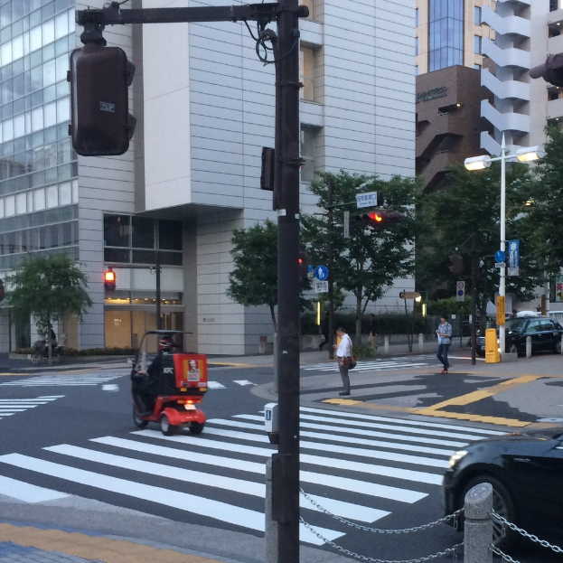 Ein Roller befindet sich auf der Straße mit einigen Personen dahinter, eine Ampel im Vordergrund und Gebäude mit Bäumen im Hintergrund.