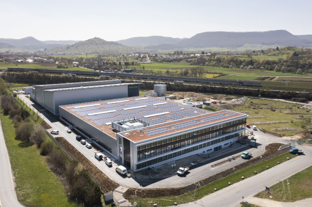 Luftaufnahme eines großeen Gebäudes mit Solarmodulen auf dem Dach, umgeben von Fahrzeugen, Bäumen, Gras und Masten, mit Höhen und einem klaren blauen Himmel im Hintergrund.
