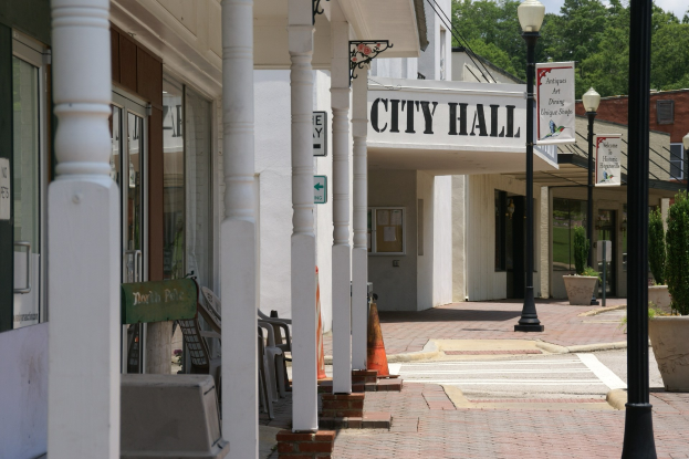 Stadtverwaltung mit umliegenden Gebäuden, Straßeninfrastruktur einschließlich Pfosten, Lampen und Namensschilder, Kraftfahrzeuge, Verkehrskegel, verstreute Stühle, Topfpflanzen, Bäume und ein bewölkter Himmel.