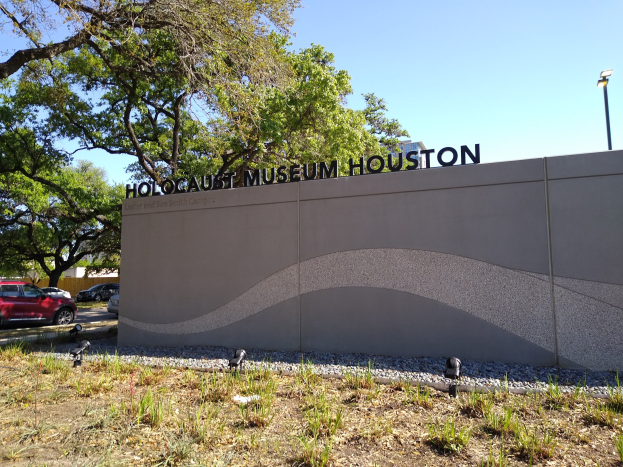 Außenansicht des Houston Holocaust Museums mit einer Textwand, Gedenksteine, Landschaftsgestaltung, Bäume, vorbeifahrende Fahrzeuge, eine Straßenlaterne und einen bewölkten Himmel.