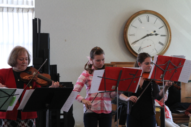 Drei Frauen auf der Bühne mit Violinen stehend und Musikständer vor sich; im Vordergrund sitzt eine Gruppe von Frauen und hört zu; an der weißen Wand hängt eine Uhr, mit einem weißen Vorhang in der linken Ecke.