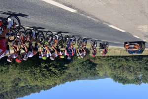 Eine Gruppe von Radfahrern mit Helmen fährt auf einer Straße während der Tour de France, mit einem Auto und Bäumen im Hintergrund und dem Himmel darüber.