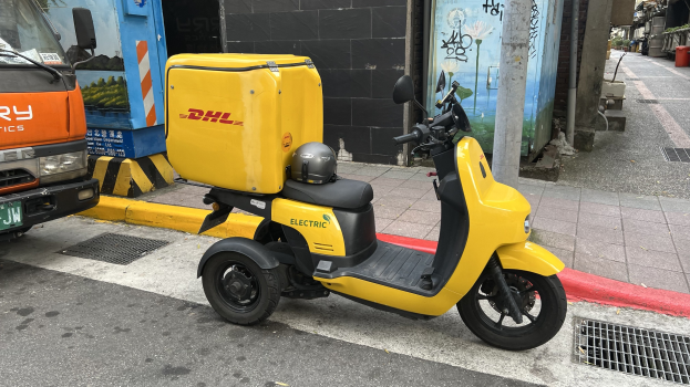 Gelber Elektroroller auf einer Straße neben einem Lastwagen mit Helm geparkt, Gebäude im Hintergrund und Menschen auf dem Gehweg.