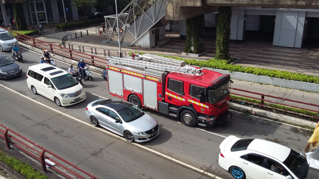 Löschfahrzeug fährt auf einer Straße mit Autos, Motorrädern und anderen Fahrzeugen; Gebäude und Bäume im Hintergrund.