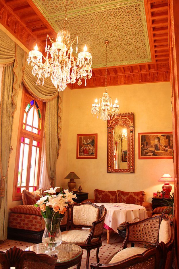 Ein Wohnzimmer mit einem Tisch und Blumentopf im Vordergrund, umgeben von Stühlen, Sofas und einem abgedeckten Tisch im Hintergrund, gerahmte Wand, hängende Lampen und ein Fenster mit Vorhängen auf der linken Seite.