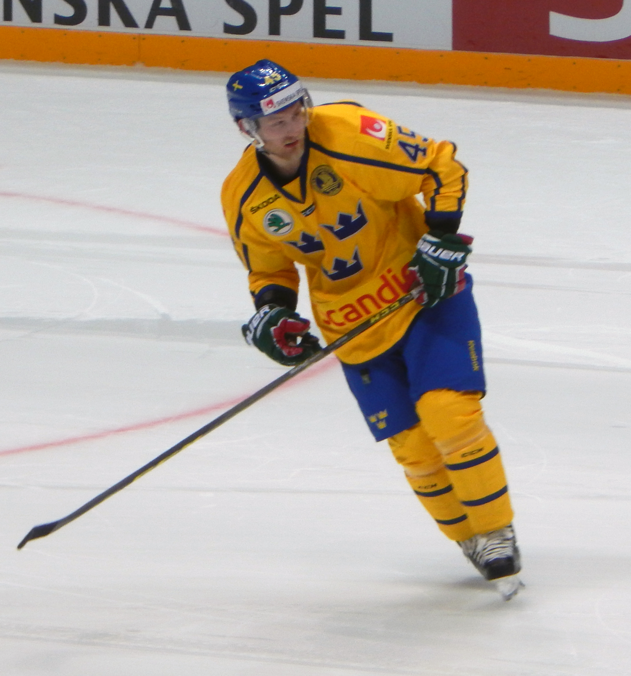 Ein Hockey-Spieler in Aktion auf dem Eis, der einen Helm, Handschuhe und Schlittschuhe trägt und einen Hockey-Schläger hält, mit einer Tafel im Hintergrund.
