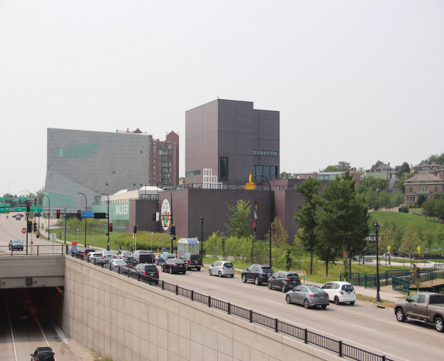 Eine Stadtstraße mit Autos, eine Brücke mit Geländern, Laternen, Verkehrsampeln, Bäumen, Gebäuden mit Fenstern und einen Himmel im Hintergrund.