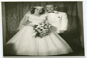 Ein Schwarz-Weiß-Foto einer Braut in einem weißen Kleid mit einem Strauß Blumen in der Hand und einem strahlenden Bräutigam neben ihr, mit einem Vorhang im Hintergrund.