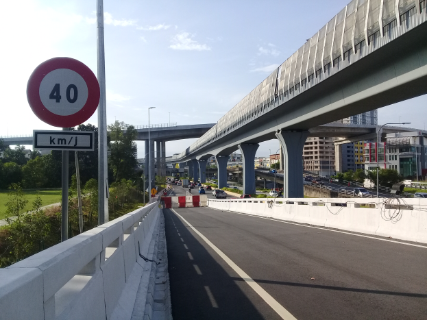 Eine Autobahn mit einem Tempolimit-Schild, Fahrzeugen, einer Brücke mit Pfeilern, Laternen, Bäumen, Gebäuden und einem bewölkten Himmel.