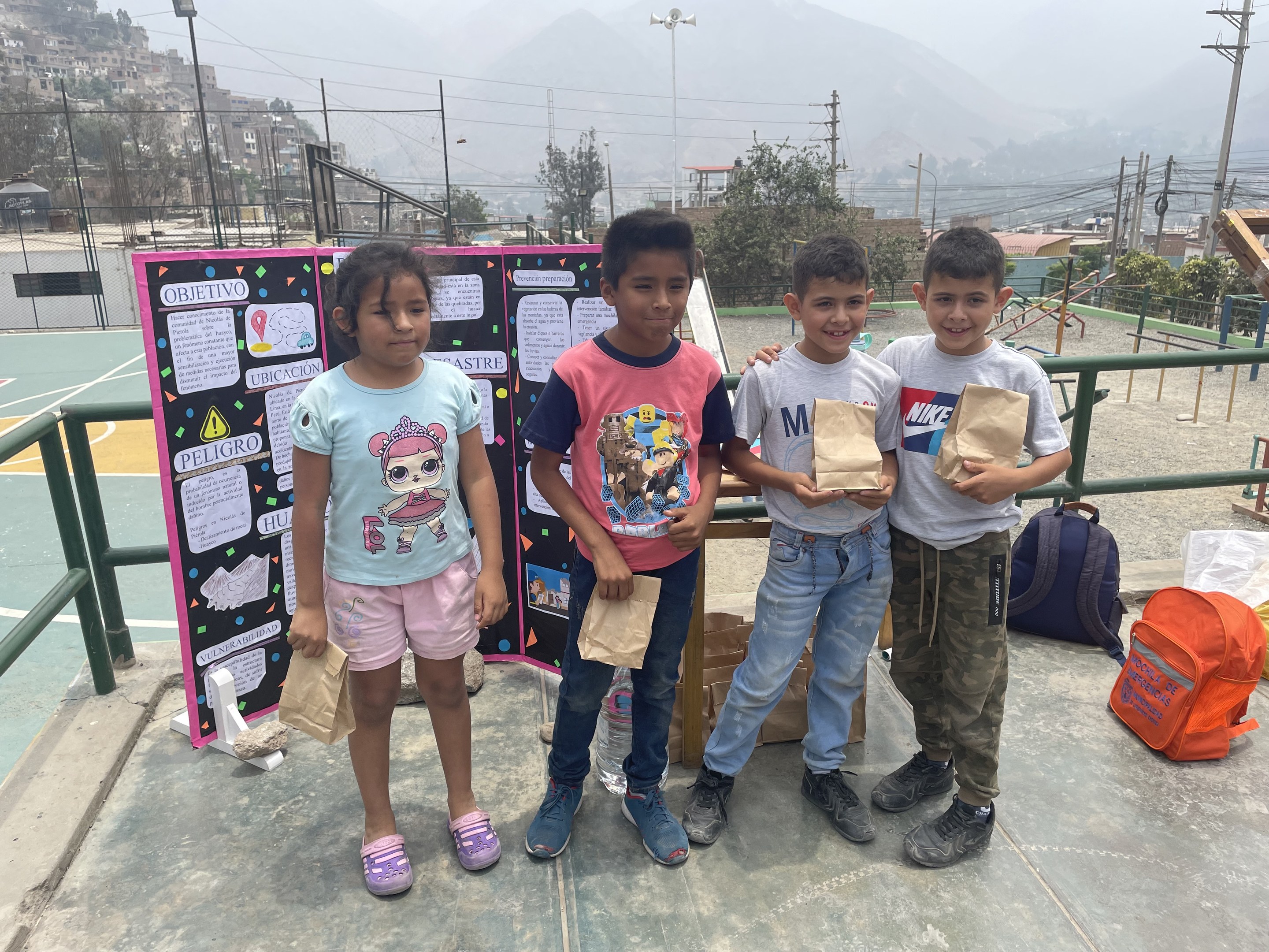 Gruppe von Kindern auf einem Basketballfeld mit Papiertüten in den Händen, Tüten und Gegenstände rechts und eine Tafel mit Text links, vor einem Hintergrund aus Zaun, Pfosten, Laternen, Bäumen, Gebäuden, Hügeln und Himmel.