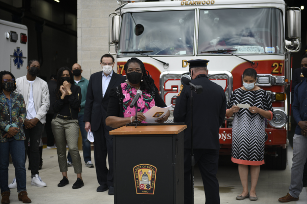 Bürgermeisterin von Chicago Lori Lightfoot steht an einem Podium mit Mikrofonen und Papieren, spricht auf einer Pressekonferenz vor einem Feuerwehrauto, umgeben von maskierten Personen, mit einer beleuchteten Wand im Hintergrund.