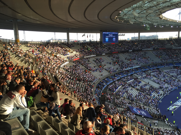 Eine große Menschenmenge sitzt im Allianz Arena Stadion in München, Deutschland, und schaut ein Fötballspiel, mit einer Bühne auf der rechten Seite, Fahnen, Masten und einem Bildschirm im Hintergrund und der Himmel oben sichtbar.