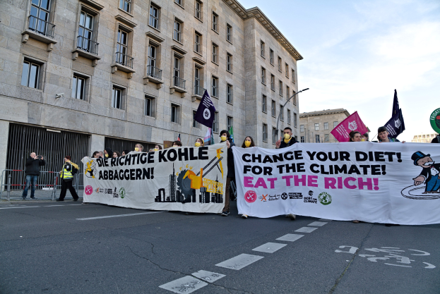 Eine Gruppe von Menschen marschiert mit einem Banner, auf dem 'Verändere deine Ernährung für das Klima, iss die Reichen' steht, eine Straße entlang. Gebäude und Laternen säumen die Straße unter einem bewölkten Himmel.