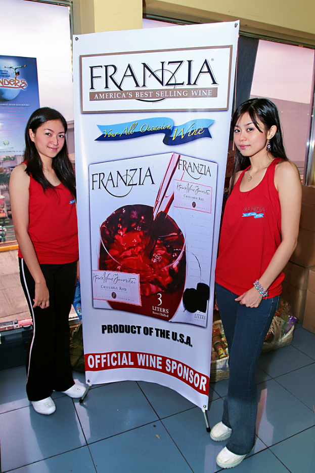 Zwei Frauen neben einem Plakat, auf dem der bestverkaufende Wein von Franzia in Amerika angekündigt wird, mit einem Glasfenster im Hintergrund.