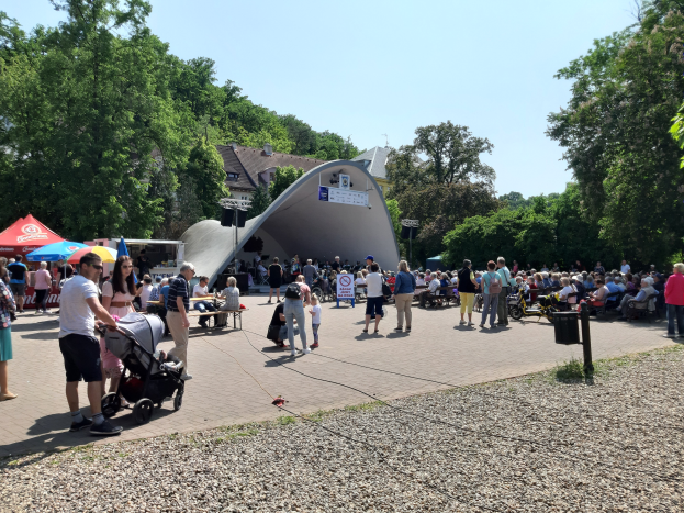 Eine Menschenmenge bei der Festivalöffnung, einige sitzen auf Bänken, andere stehen, mit einem Kinderwagen im Vordergrund, Zelten links, einem Müllcontainer rechts und Gebäuden und Bäumen im Hintergrund unter einem klaren blauen Himmel.
