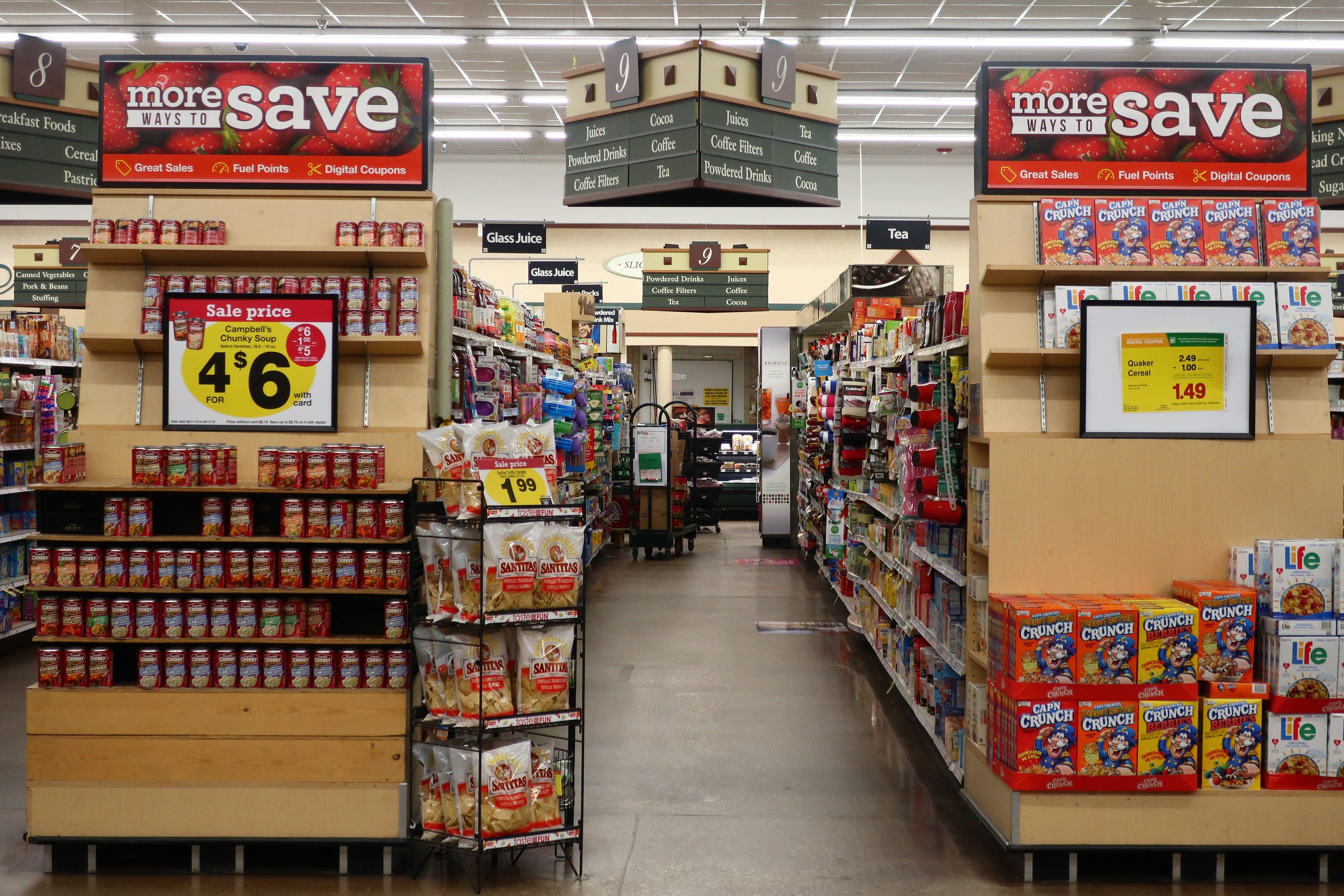 Ein Innenraum eines Lebensmittelladens mit Regalen und Boden voller verschiedener Lebensmittel, Deckenbeleuchtung, Informationsschildern und einer Person im Hintergrund.