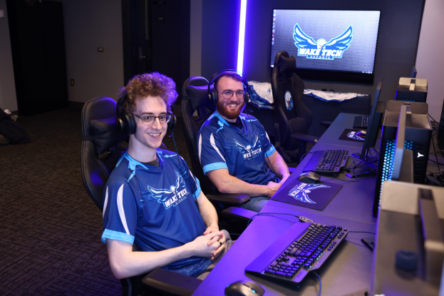 Zwei Männer in Headsets lächeln, während sie an einem Schreibtisch mit Monitoren, Tastaturen und anderem Equipment sitzen und scheinbar ein Spiel in einem Raum mit einer wandmontierten Bildschirm und Bodenunordnung spielen.