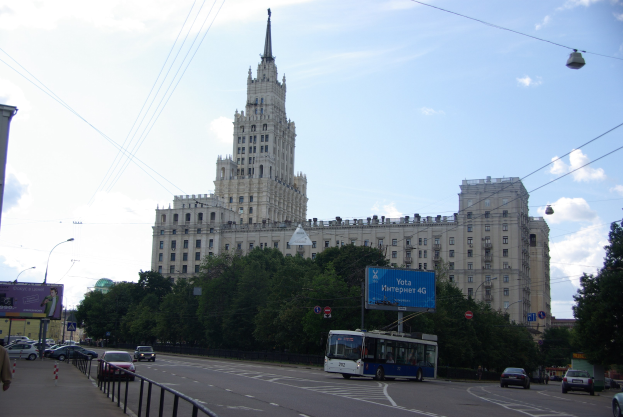 Eine belebte Straße in Moskau, Russland, mit Fahrzeugen, einem Fußgänger, Geländern, Bäumen, Laternen, Strommasten mit Drähten und einem großen Gebäude mit einem Uhrenturm im Hintergrund unter einem bewölkten Himmel.