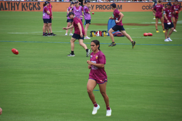 Frauen beim Australian-Rules-Fußball auf einem grünen Feld, mit verstreuten Bällen und einem Banner im Hintergrund, einige tragen Kappen und Turnschuhe.