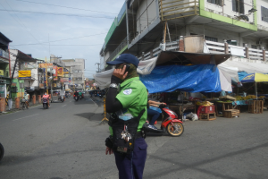 Ein Polizist in grüner und blauer Uniform mit blauer Mütze steht auf einer Straße, spricht in ein Handy, mit Fahrzeugen auf der Straße hinter ihm und Marktbuden zu seiner Rechten, unter einem bewölktem Himmel.