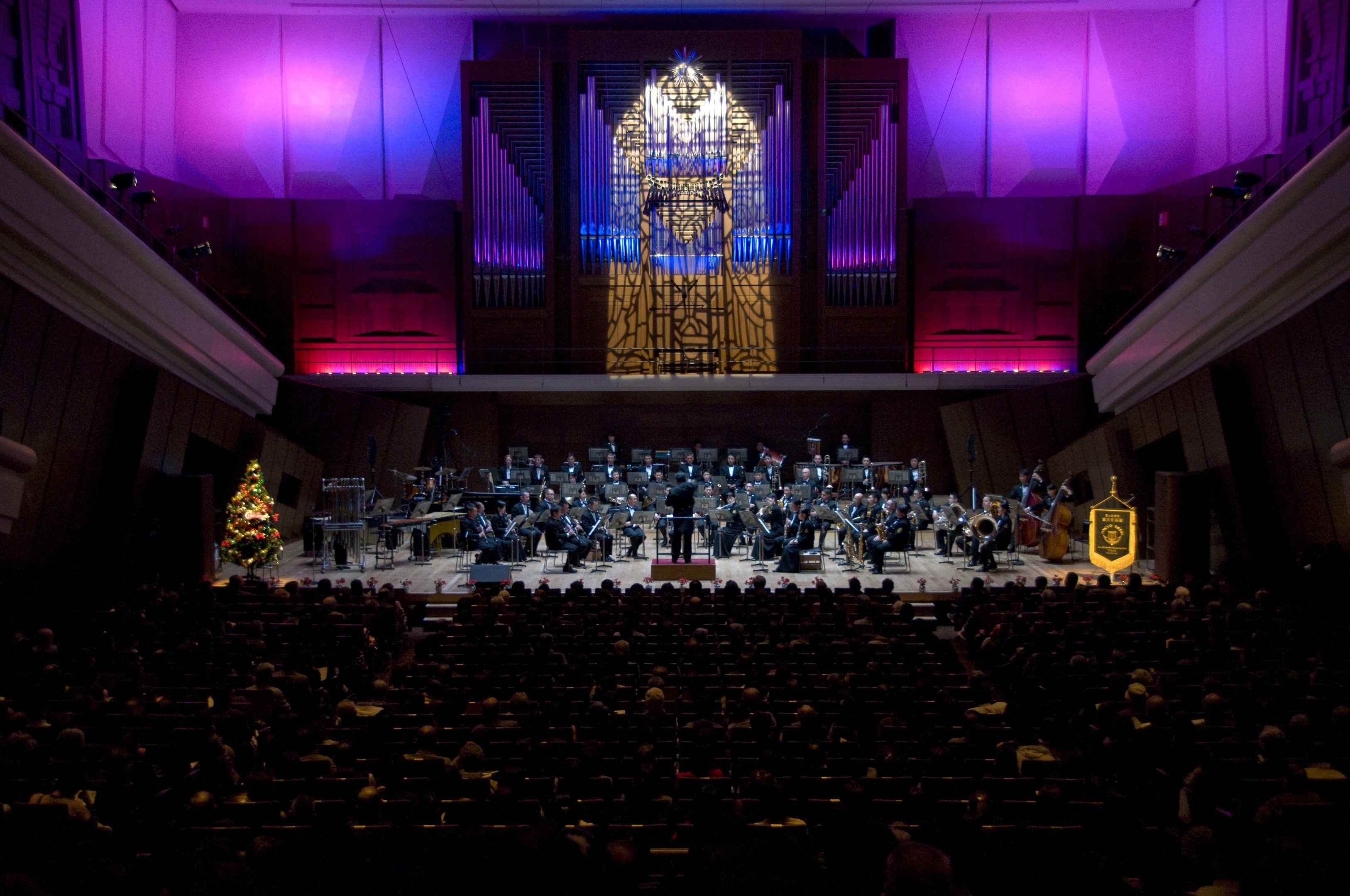 Großer Saal mit Publikum bei einem Konzert; Musiker auf der Bühne mit Instrumenten und sitzenden Musikern; dekorierter Weihnachtsbaum und beleuchtete Wände im Hintergrund.