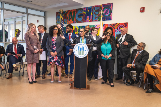 Gruppe von Menschen vor einem Rednerpult mit Mikrofon und einer Tafel mit der Aufschrift "Sancho Stadtverordneter tritt dem Stadtrat bei", mit sitzenden und stehenden G├Ąsten, Flaggen, Feuerl├Âscher und Deckenleuchten.