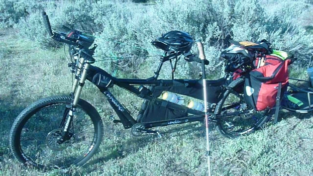 Ein Tandemfahrrad mit Gep√§ck und Anh√§nger, das auf einer grasbewachsenen Fl√§che positioniert ist.