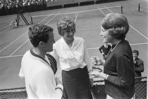 Schwarz-weißes Foto von zwei Frauen und einem Mann auf einem Tennisplatz, wobei eine Frau eine Brille und der Mann ein Objekt hält, und ein Zaun und Zuschauer im Hintergrund.