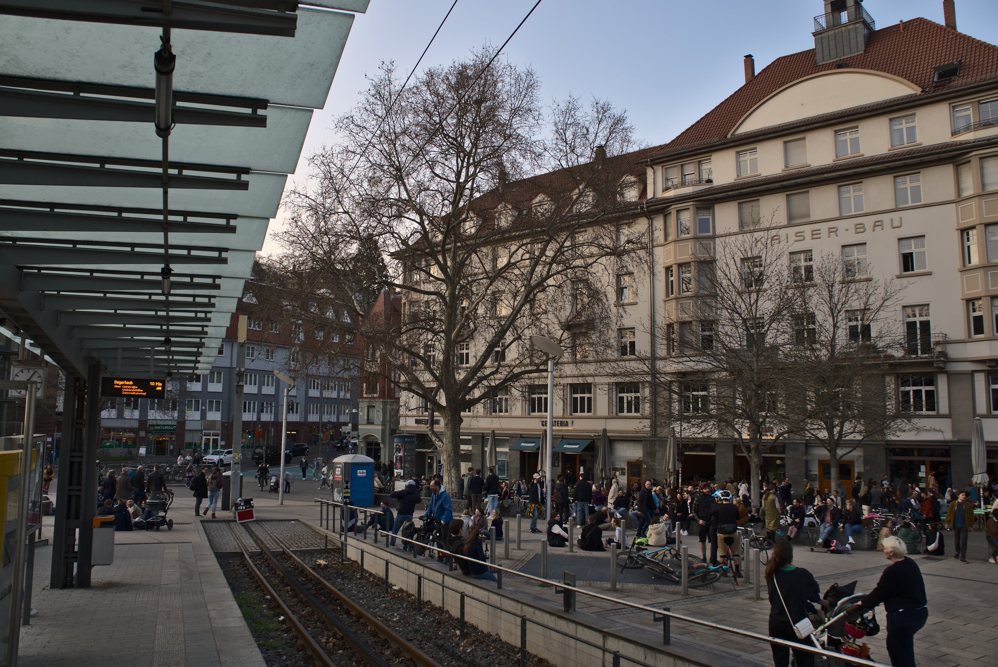 Eine belebte Stadtstraße mit einem Bahnhof auf der linken Seite, Menschen gehen, sitzen auf Bänken und stehen, umgeben von Gebäuden, Bäumen, Laternen und Texttafeln unter einem sichtbaren Himmel.