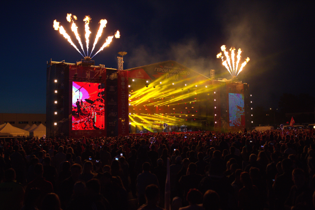 Eine große Menschenmenge versammelte sich auf einem Musikfestival mit einer Bühne in der Mitte, auf der Musiker im Scheinwerferlicht spielten und von Fahnen umgeben waren, Zelte links und ein dunkler Himmel mit Feuerwerk im Hintergrund.
