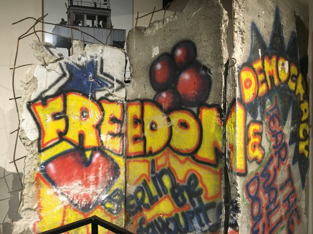 Eine Wand mit kühner, schwarzer Graffiti, die 'Freiheit und Demokratie' lautet, in einem Raum mit einem Geländer unten und einem Gebäude mit Fenstern im Hintergrund.