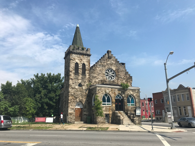 Eine alte steinerne Kirche mit einem Kirchturm an der Kreuzung Main Street und Main Street, umgeben von Gebäuden, Straßenlaternen, Straßenlaternen, Kraftfahrzeugen, Fußgängern, Werbetafeln, Bäumen und einem bewölkten Himmel.