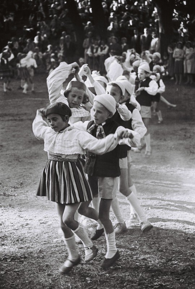 Schwarß-weißes Foto von Kindern, die in einem Park mit Bäumen und einigen stehenden Beobachtern im Hintergrund tanzen.