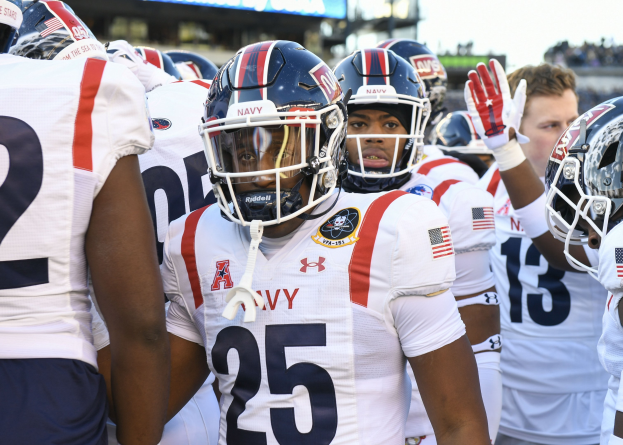 Gruppe von Männern in Football-Uniformen auf einem Feld mit Helmen und Handschuhen, mit einer unscharfen Menge, Anzeigetafel mit 'Auburn vs Virginia Tech - 11/28/15 College Football Pick, Odds, and Prediction' und Himmel im Hintergrund.