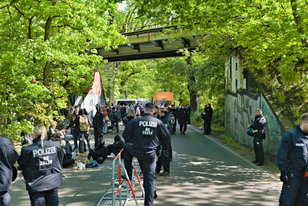 Polizeibeamte hinter einer Barriere auf einer Straße mit sitzenden und stehenden Personen, Bäumen, einer Brücke und einer Wand mit Schrift im Hintergrund.