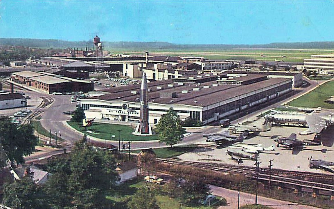 Altes Schwarz-Weiß-Foto eines Flughafens mit geparkten Flugzeugen, Gebäuden, Bäumen, Straßen, Fahrzeugen, Strommasten, Gras, Bahnschienen, Türmen, Hügeln und einem klaren blauen Himmel; Text erscheint unten.