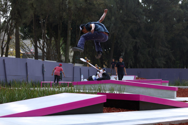 Ein Skateboarder führt einen Sprung auf einer Rampe aus, während Zuschauer, einige mit Skateboards, in der Nähe stehen; es sind auch Taschen, Flaschen und andere Gegenstände verstreut, Gras, eine Wand, Bäume, Gebäude und ein klarer blauer Himmel im Hintergrund zu sehen.