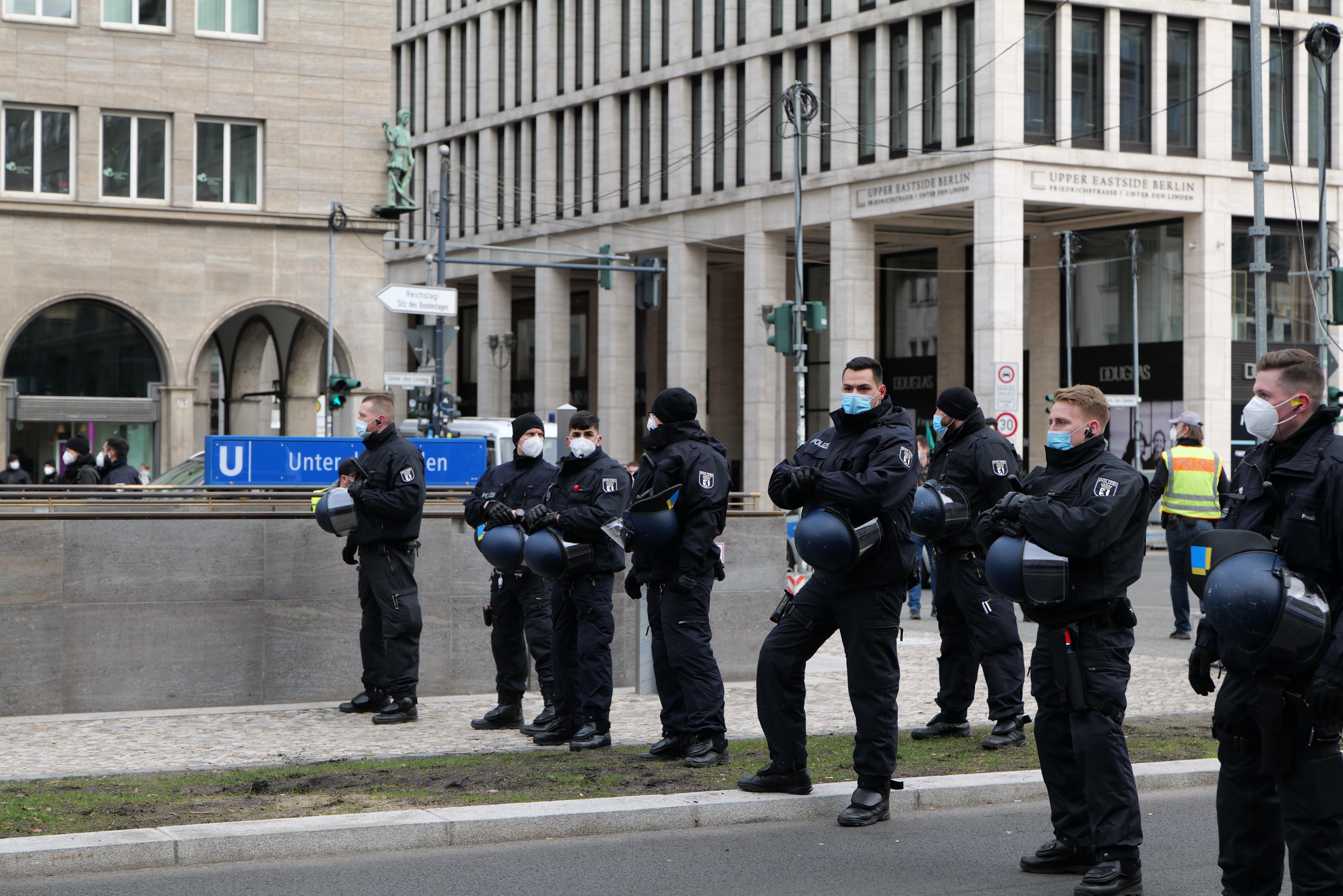 Eine Gruppe von Polizisten in schwarzen Uniformen und Masken steht vor einem Berliner Gebäude mit Glasfenstern und Säulen, einige halten Helme, mit Laternenmasten, Verkehrsampeln, Schildern mit Text, einer Statue und anderen Menschen im Hintergrund auf einer grünen Fläche.