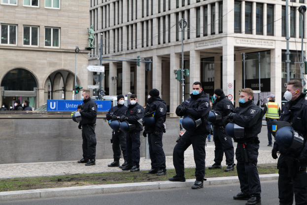 Eine Gruppe von Polizisten in schwarzen Uniformen und Masken steht vor einem Berliner Gebäude mit Glasfenstern und Säulen, einige halten Helme, mit Laternenmasten, Verkehrsampeln, Schildern mit Text, einer Statue und anderen Menschen im Hintergrund auf einer grünen Fläche.