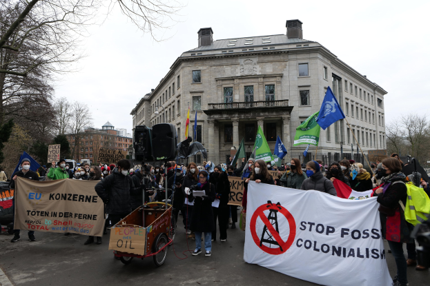 Eine große Gruppe von Menschen marschiert in einem Protest gegen fossile Brennstoffe, trägt Schilder und Fahnen, mit einem Fahrzeug im Vordergrund und Gebäuden, Bäumen und einem klaren blauen Himmel im Hintergrund.