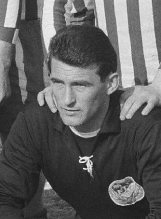 Ein Schwarz-Weiß-Foto eines Mannes, der auf einem grünen Fußballfeld sitzt und ein T-Shirt und eine Kette trägt, während zwei Personen hinter ihm stehen.